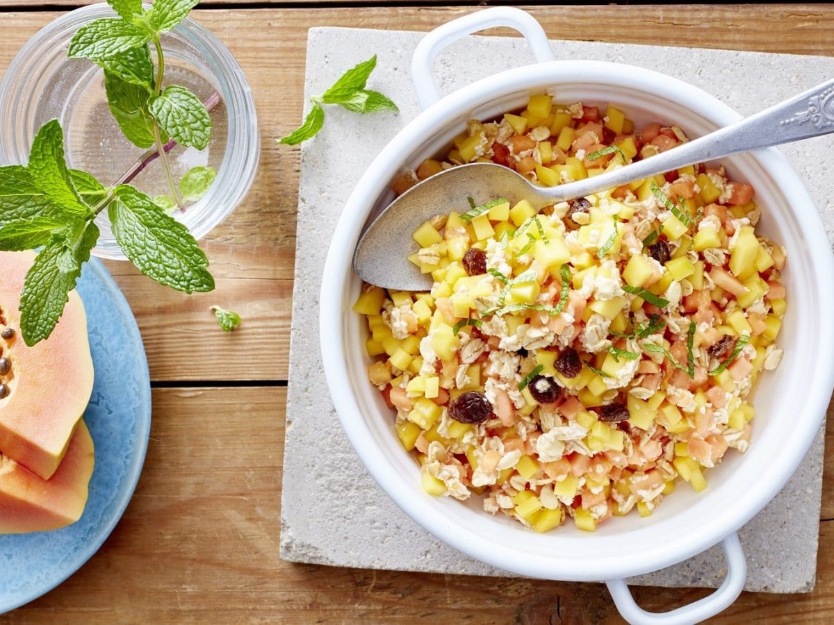 Exotisches Müsli mit Mango, Papaya, Nüssen, Rosinen und Kokosflocken, garniert mit frischer Pfefferminze und serviert in einer weißen Schale auf einem klassischen Holztisch