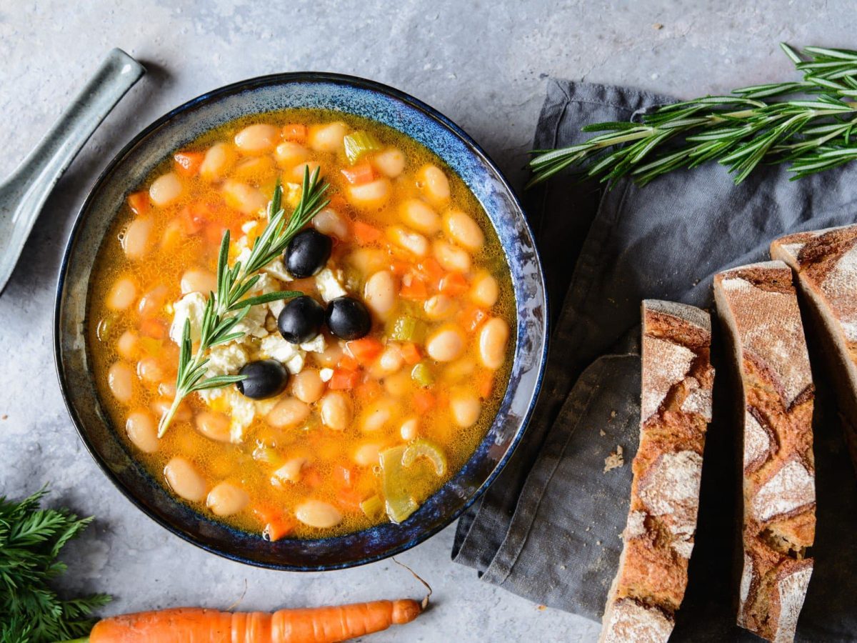 Eine dunkelblaue Suppenschale mit der Bohnensuppe auf grauem Tisch mit Bundmöhren und Landbrot an der Seite.