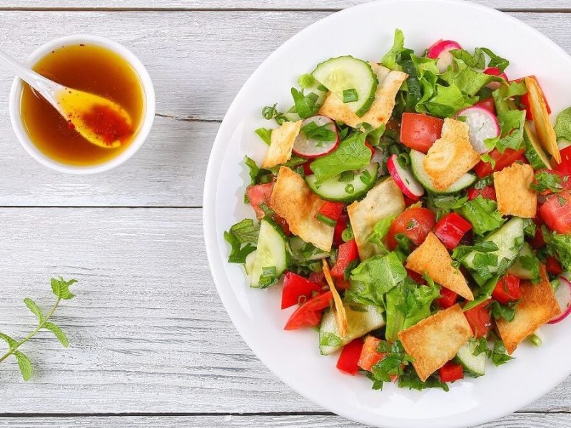 Eine weiße Schüssel mit Fattoush steht auf einem weißen Holztisch.