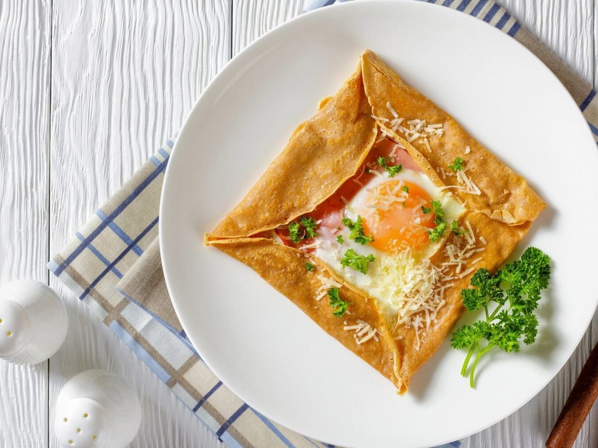 Auf einem weißen Teller liegt ein Buchweizen-Buchweizen-Pfannkuchen mit Schinken, Ei und Käse