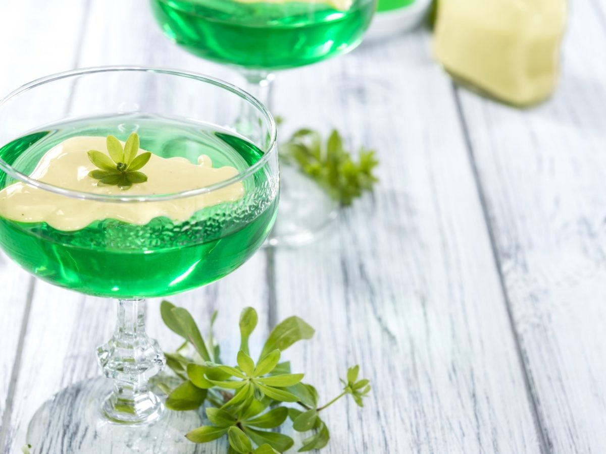 Zwei Gläser mit grüner Götterspeise und Vanillesauce auf einem weißen Holztisch mit frischem Waldmeister.