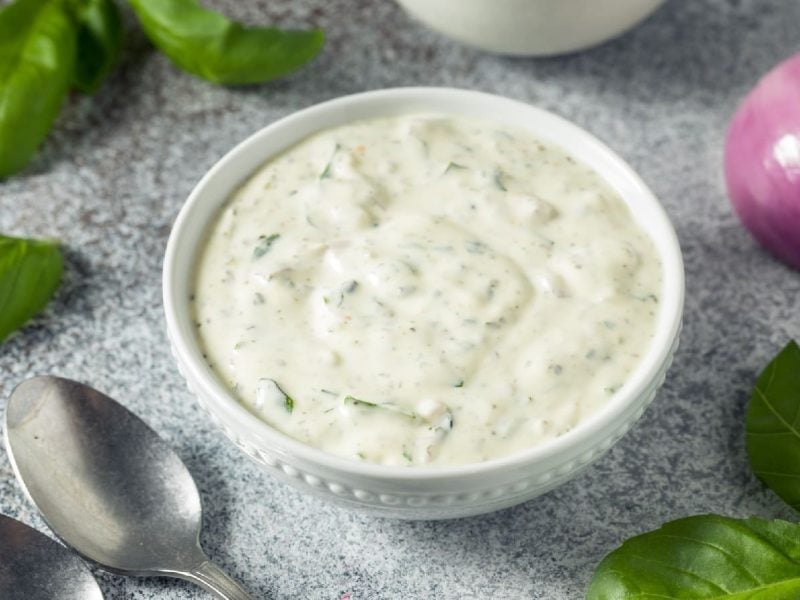 Green Goddess Dip vin einem weißen Gefäß neben zwei Löffeln und Basilikumblättern