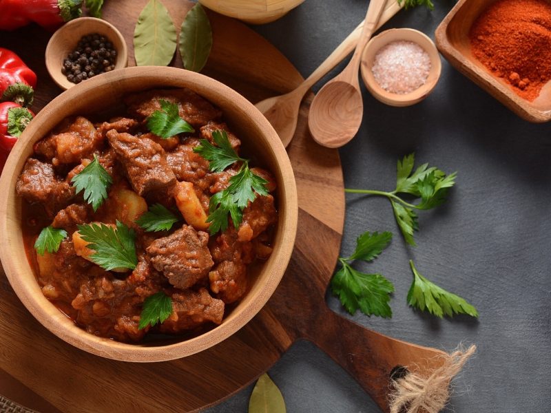 Eine Holzschale mit Gulasch und allerhand Gewürzen und Kräutern auf einem grauem Tisch.