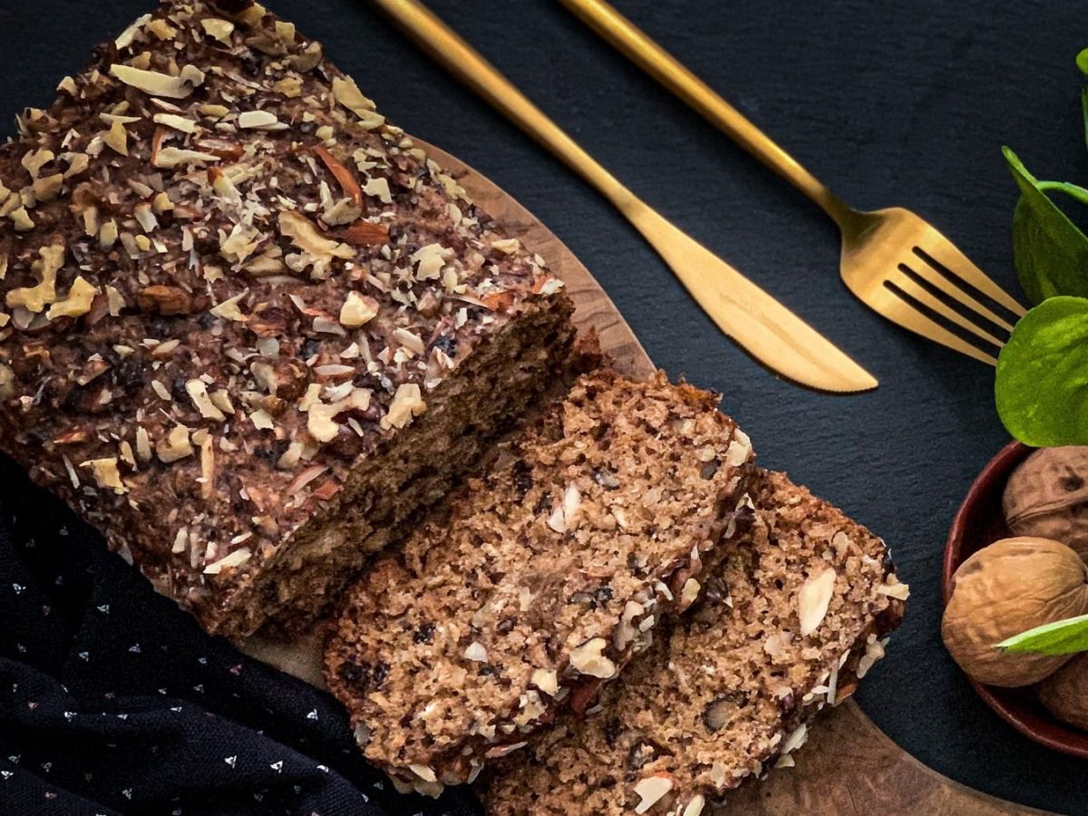 Ein angeschnittenes Haferflocken-Brot ohne Backen in der Draufsicht, daneben liegt goldenen Besteck, außerdem sind Walnüsse in einer Schale zu sehen.