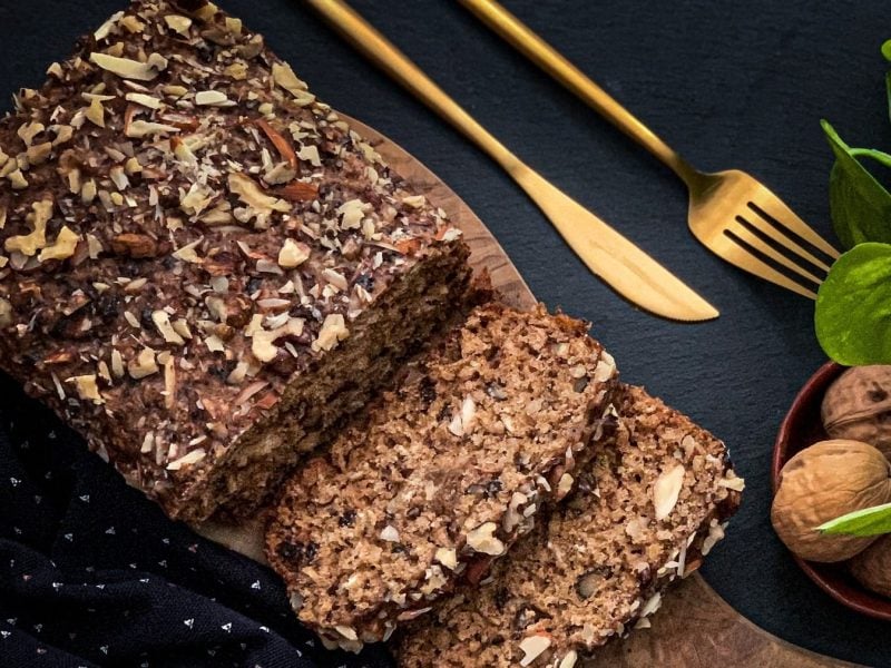 Ein angeschnittenes Haferflocken-Brot ohne Backen in der Draufsicht, daneben liegt goldenen Besteck, außerdem sind Walnüsse in einer Schale zu sehen.