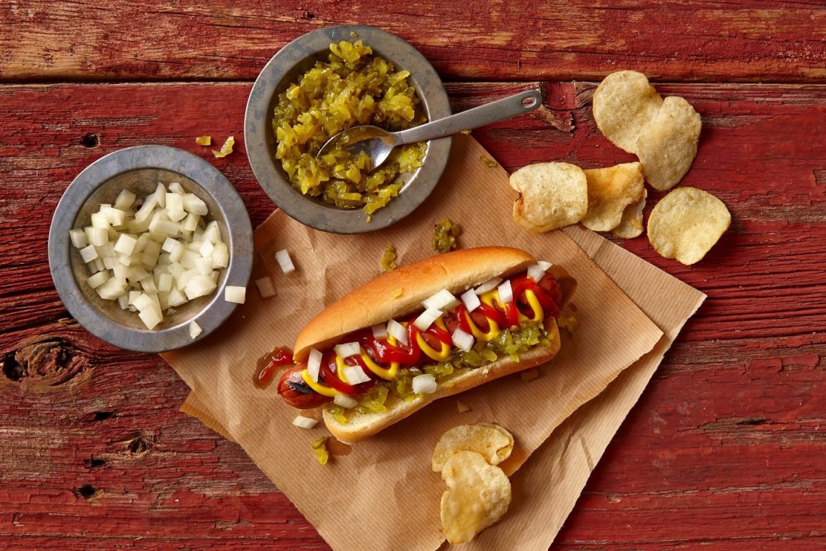 Woher kommt der Name Hotdog? Dazu ein Hotdog auf Backpapier mit Kartoffelchips. Daneben je eine graue Schale mit gewürfelten Zwiebeln und sauren Gurken auf rotem Holztisch. Aufnahme aus der Vogelperspektive.