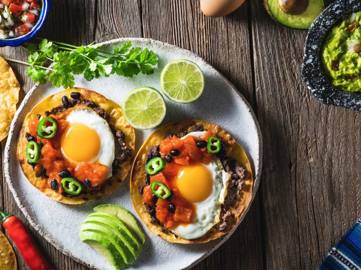 Huevos ranchero mexikanisches Frühstück auf Teller mit Limette und Avocado. Daneben Tortilla, Salsa und Guacamole. Chilis, Avocados und Tomaten als Dekoration im Bild. Arrangiert auf Holztisch. Aufnahme aus der Vogelperspektive.