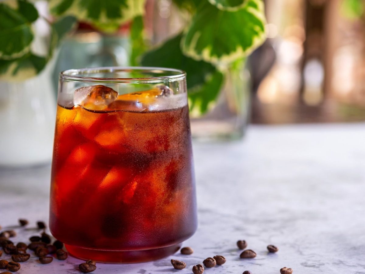 Ein Glas Iced Americano mit Eiswürfeln, darum herum liegen Kaffeebohnen, im Hintergrund stehen Pflanzen.