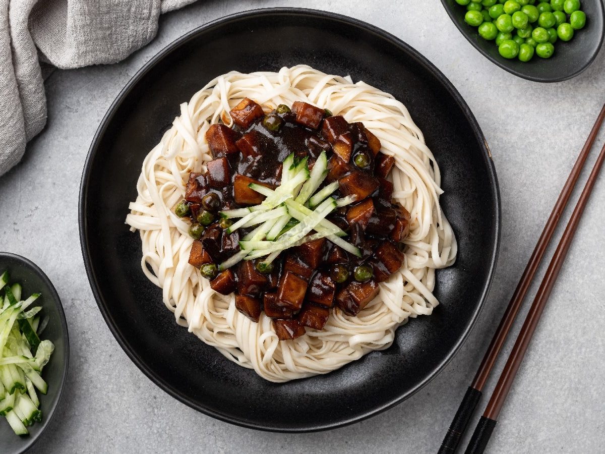 Eine Schüssel Jajangmyeon auf einem grauen Tisch neben einer Schale Gurkenstreifen und Erbsen