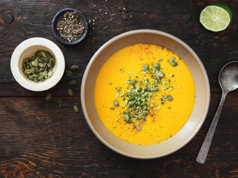 Kalte Gemüsesuppe mit Gurken, Möhren und Paprika, bestreut mit Körnern und Samen, auf dunklem Untergrund in der Draufsicht, daneben stehen einige Zutaten.