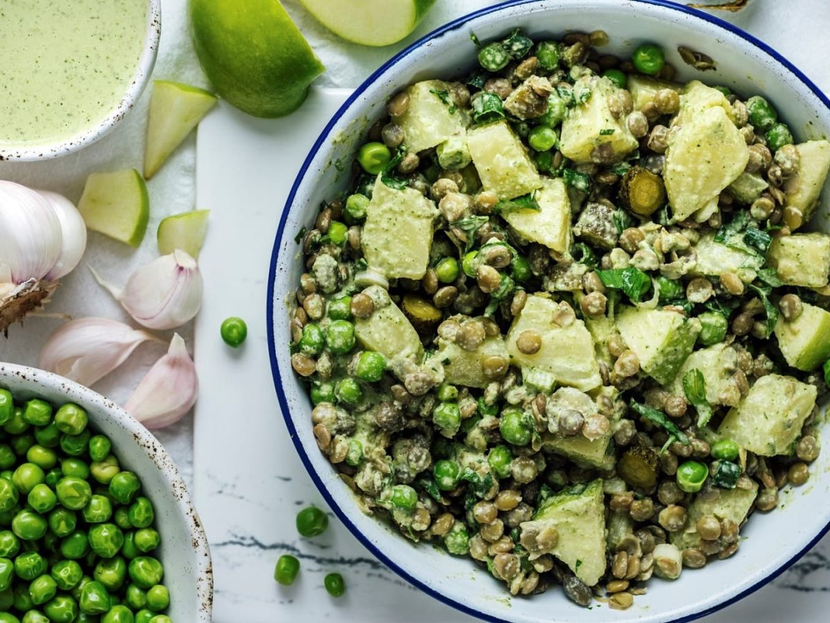 eine Schale veganer Kartoffelsalat mit Äpfeln und Erbsen in der Draufsicht, daneben sind Zutaten wie Mayo, Erbsen und Frühlingszwiebeln zu sehen.