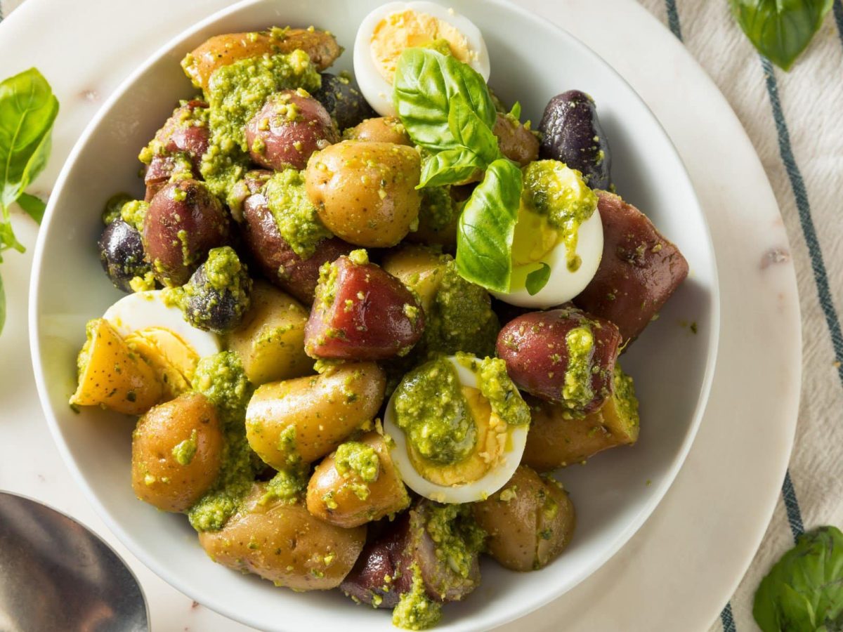 Kartoffelsalat mit Pesto, Kalamata Oliven und Ei in weißer Schale auf weißen Teller auf blau-weißem Tischtuch. Basilikumblätter als Deko auf dem Salat und daneben im Bild. Metalllöffel unten links erkennbar. Aufnahme aus der Vogelperspektive.