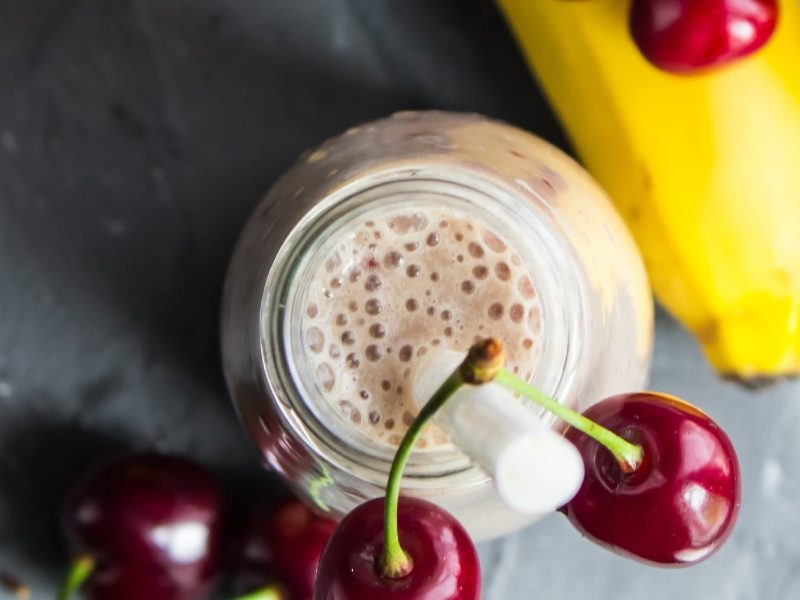 Ein Milkshake von oben fotografiert mit Kirschen und Bananen drum herum auf einem grauen Untergrund.