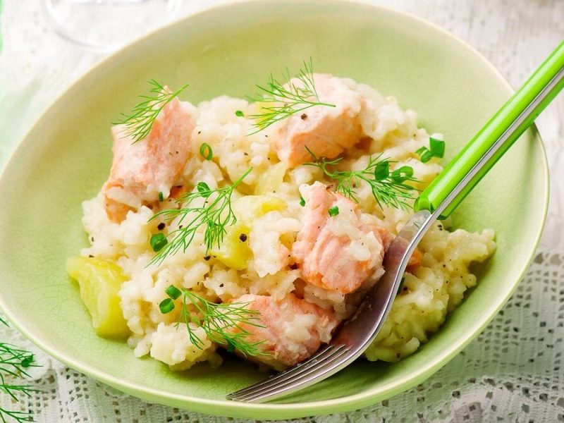 Auf einem hellen Tisch steht eine grüne Schüssel mit einem Lachs-Risotto mit Sekt. In der Risotto steckt ein grüner Löffel.