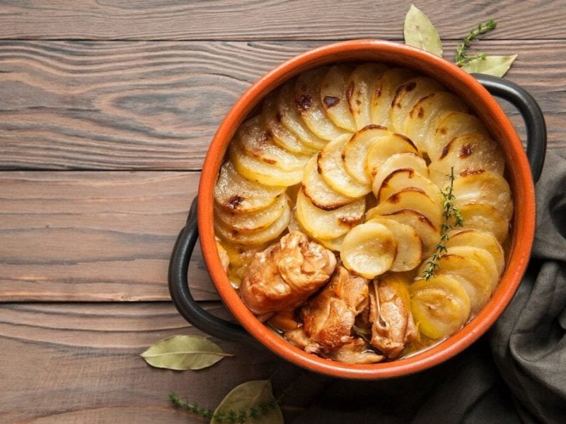 Eine Schüssel aus Ton mit einem Lancashire-Hotpot steht auf einem Untergund aus Holz. Daneben liegen Lorbeerblätter und ein Tuch.