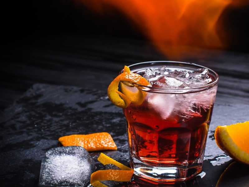 Mezzoni Cocktail mit Mezcal wie Negroni mit Eis und Orangenschale auf schwarzem Untergrund. Einswürfel und Orange als Dekoration im Bild. Flammen über dem Shortdrink. Frontalansicht.