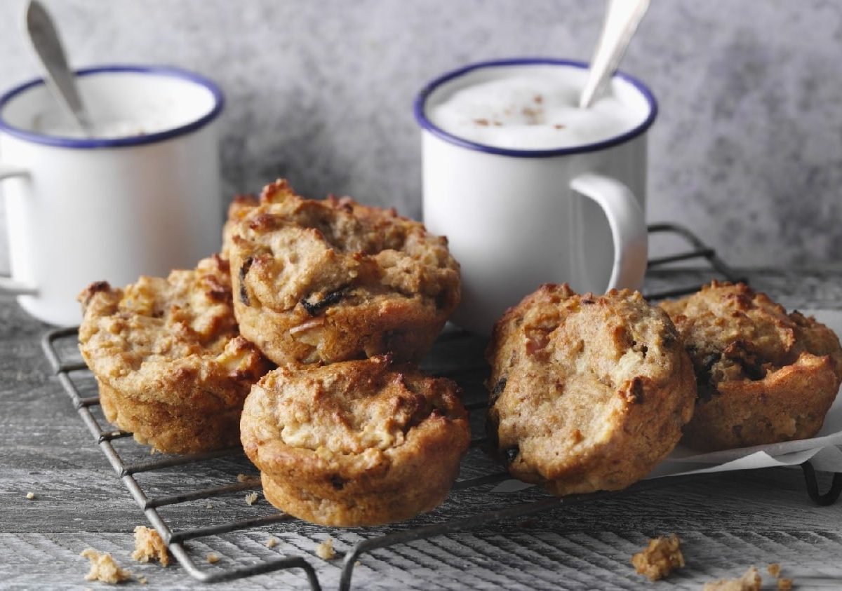 Fünf gebackene Müsli-Muffins auf einem Gitter und zwei helle Kaffeetassen im Hintergrund. Alles auf einem Holztisch und vor grauer Wand.