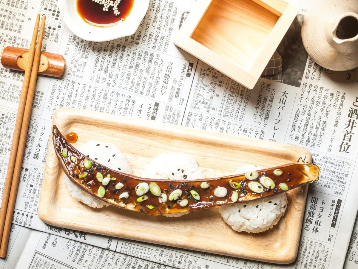 Nasu Dengaku japanische Aubergine mit Miso auf Holzbrett mit Reis und Sesam. Daneben Holzessstäbchen auf Stäbchenhalter. Oben Ponzu-Sauce und Holzschälchen und Sake. Angerichtet auf japanischer Zeitung. Aufnahme aus der Vogelperspektive.