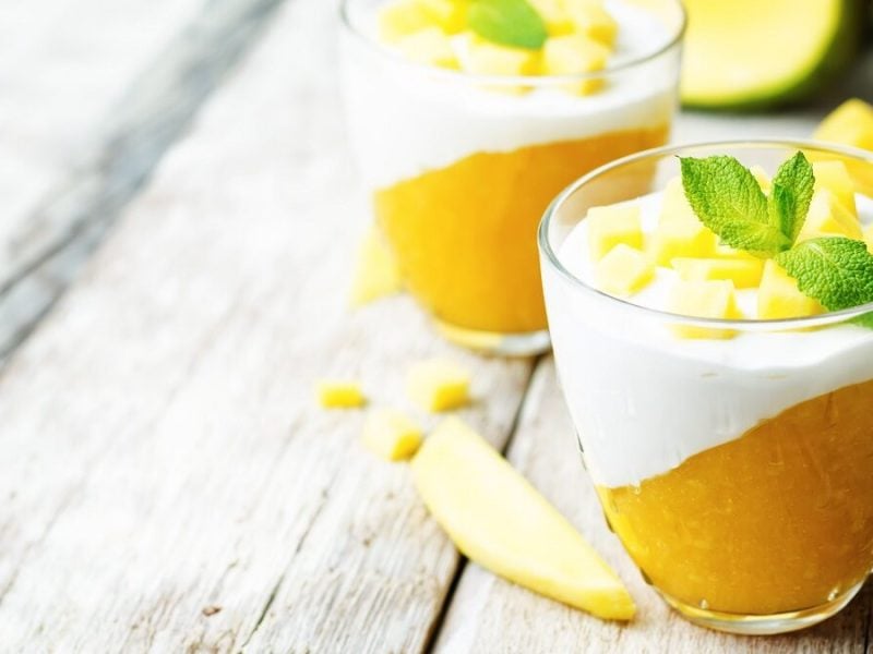 Zwei Gläser Panna Cotta mit Mango und Minze stehen auf einem Untergrund aus Holz. Daneben liegen Mango-Stücke.