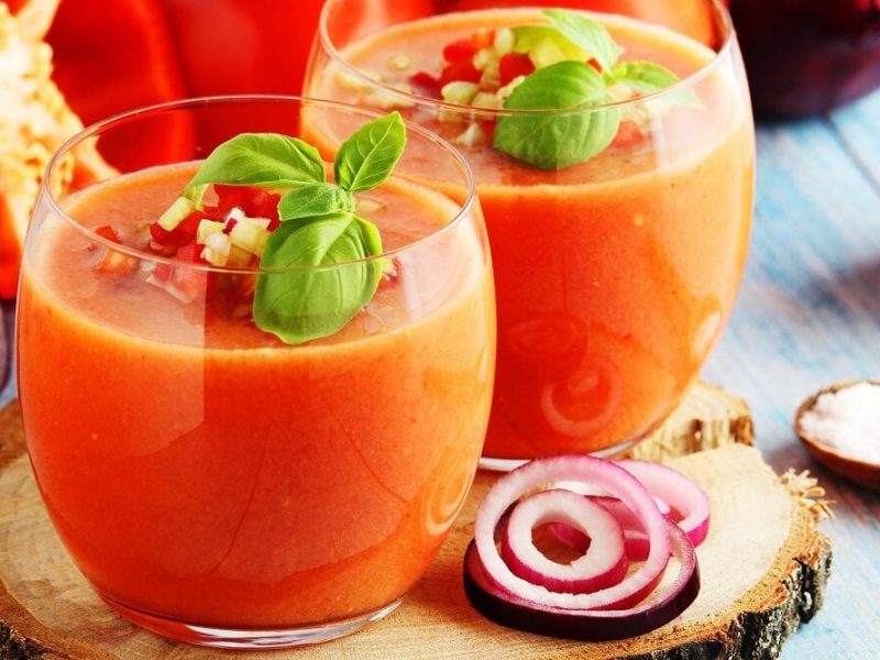 Zwei Gläser mit Paprika-Gazpacho mit Nektarine stehen auf einem Tisch. Daneben liegt ein aufgeschnittene rote Zwiebel., Und auch Sellerie liegt neben den Gläsern.