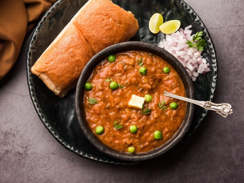 Pav Bhaji, indisches Streetfood, in einer Schale, daneben ein Milchbrötchen, rote Zwiebeln und Limettenspalten. Alles in der Draufsicht.