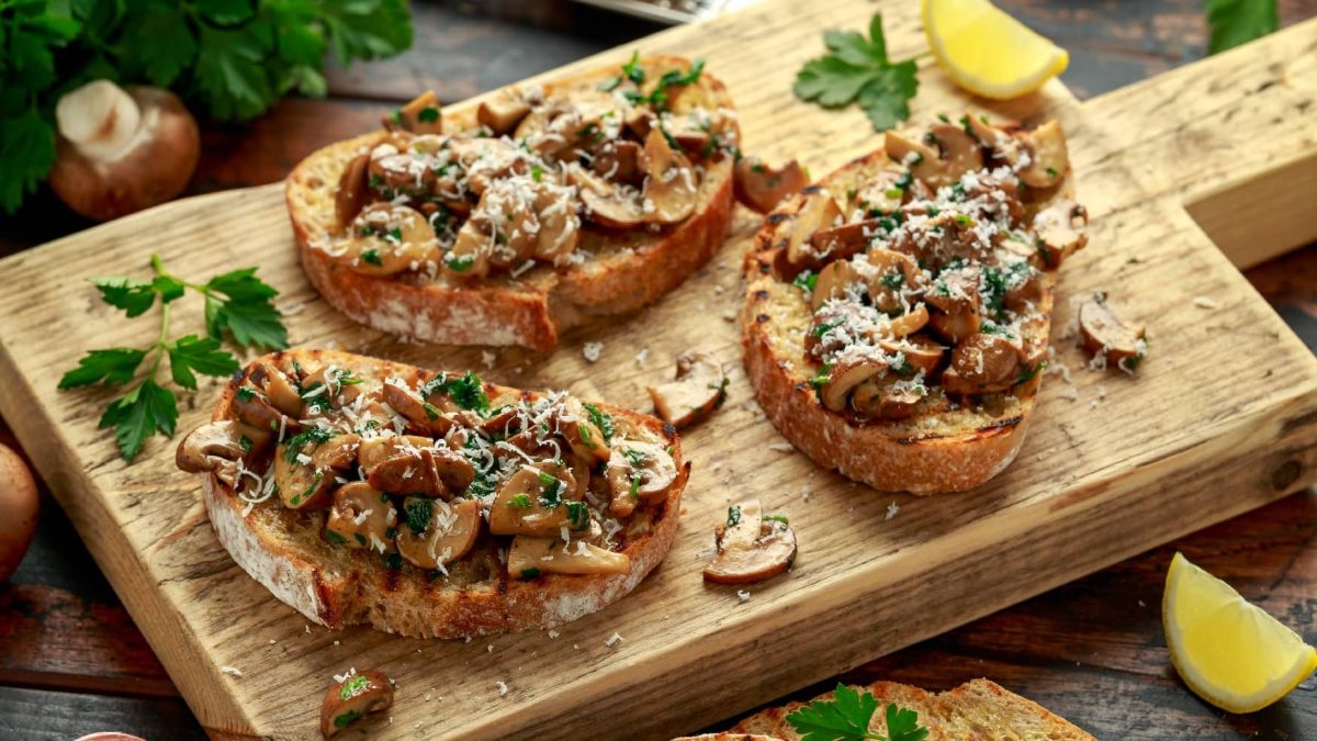 Drei Pilz-Bruschetta auf einem Holzbrett mit Parmesan und Zitronenecken. Auf dem Holztisch liegen weitere Brotscheiben Pilze und etwas Petersilie.