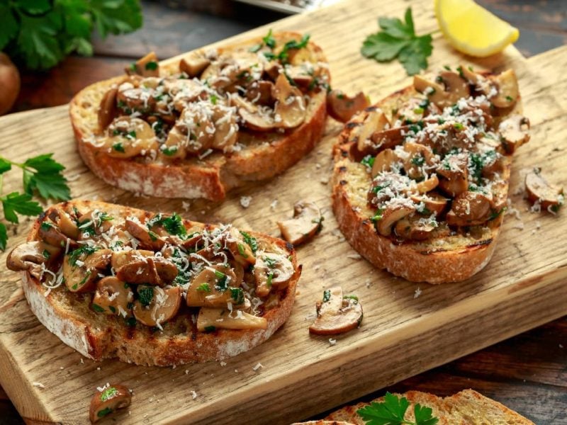 Drei Pilz-Bruschetta auf einem Holzbrett mit Parmesan und Zitronenecken. Auf dem Holztisch liegen weitere Brotscheiben Pilze und etwas Petersilie.