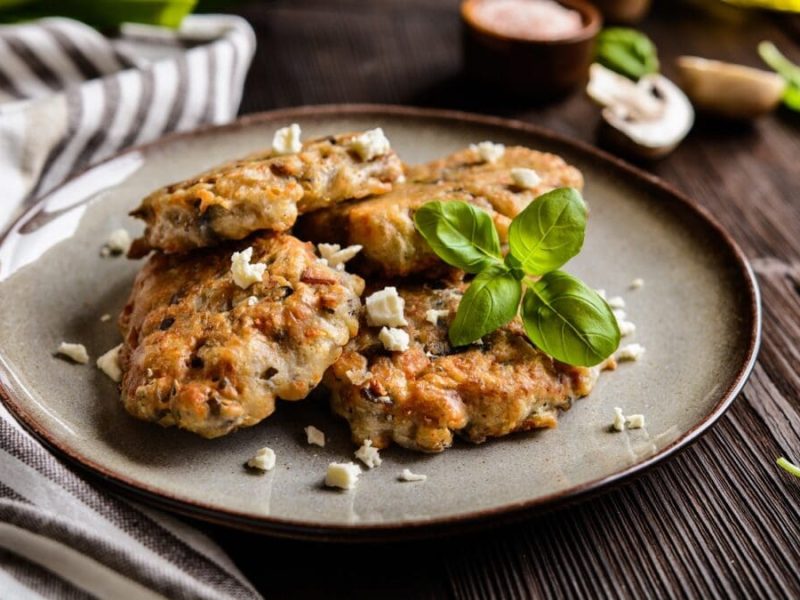 Vegetarischer Pilz-Patty mit Feta und Shiitake auf grauem Teller auf Holztisch. Basilikum und Schälchen mit Saucen im Bild. Links weiß-grau gestreiftes Stofftuch. Frontalansicht.