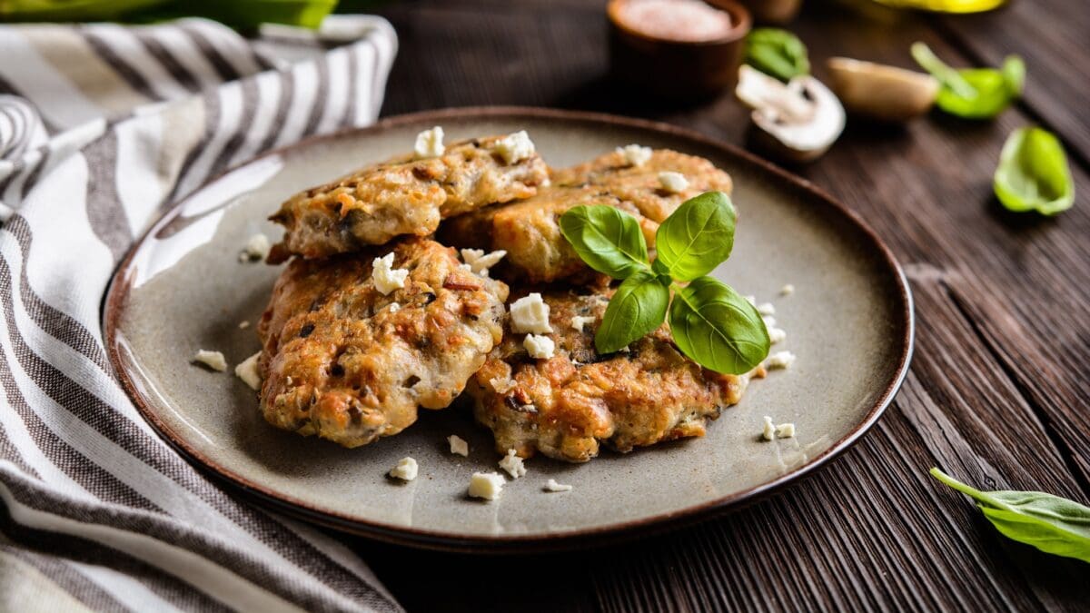 Vegetarischer Pilz-Patty mit Feta und Shiitake auf grauem Teller auf Holztisch. Basilikum und Schälchen mit Saucen im Bild. Links weiß-grau gestreiftes Stofftuch. Frontalansicht.