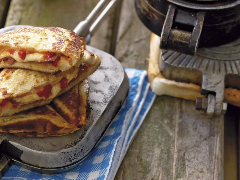 Pizza Jaffles auf einem gusseisernen Waffeleisen. Das Ganze liegt auf einem blauen Küchentuch auf einem hölzernen Untergrund.