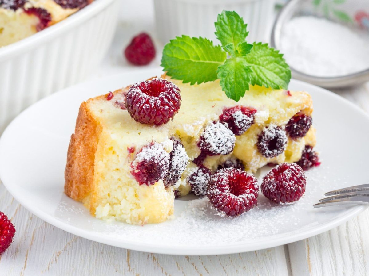 Quark-Vanille-Auflauf mit Himbeeren auf einem weißen Teller. Daneben eine Gabel und ein Sieb mit Puderzucker.