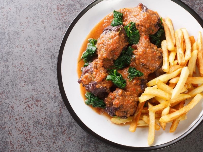 Rabo de Toro Cordoba spanischer Ochsenschwanz mit Spinat und Pommes auf Teller auf grauem Untergrund. Aufnahme aus der Vogelperspektive.