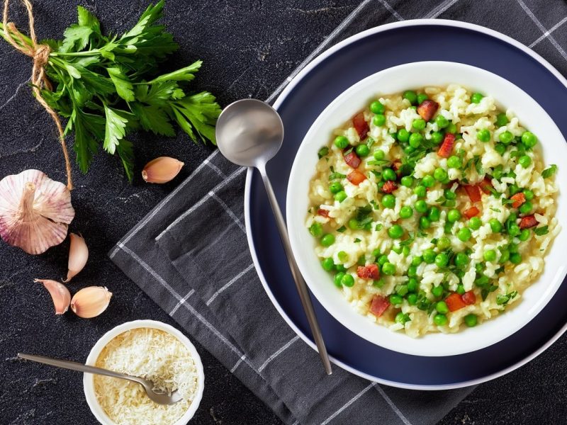 Risi Bisi 80er Retro Rezept in weißem Teller auf dunklem Holz auf grauem Untergrund. Daneben Parmesan in Schüssel, Knoblauch und frische Petersilie. Aufnahme aus der Vogelperspektive