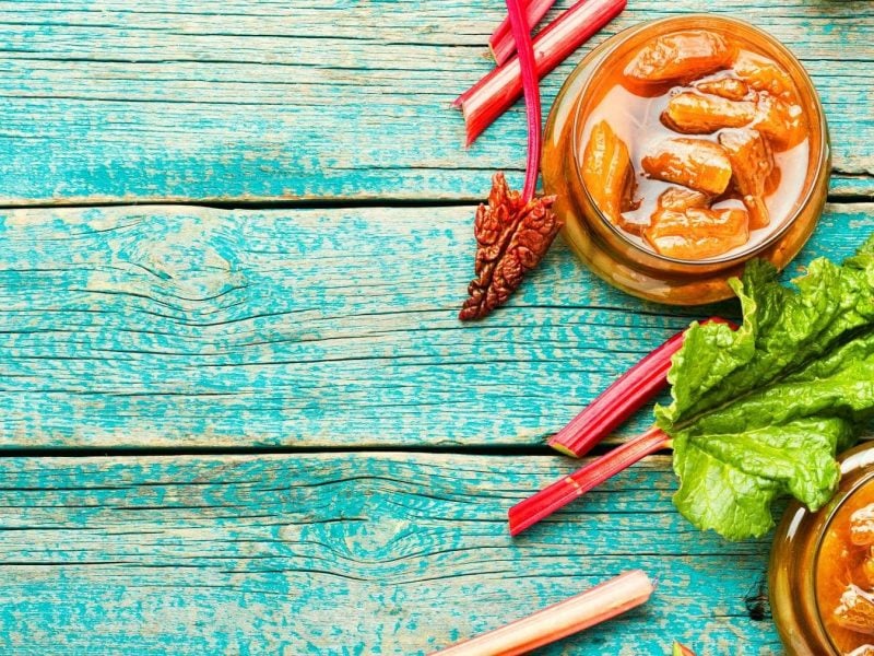 Zwei Gläser Rhabarber-Chutney mit Zwiebeln auf blauem Holz, daneben frischer Rhabarber.