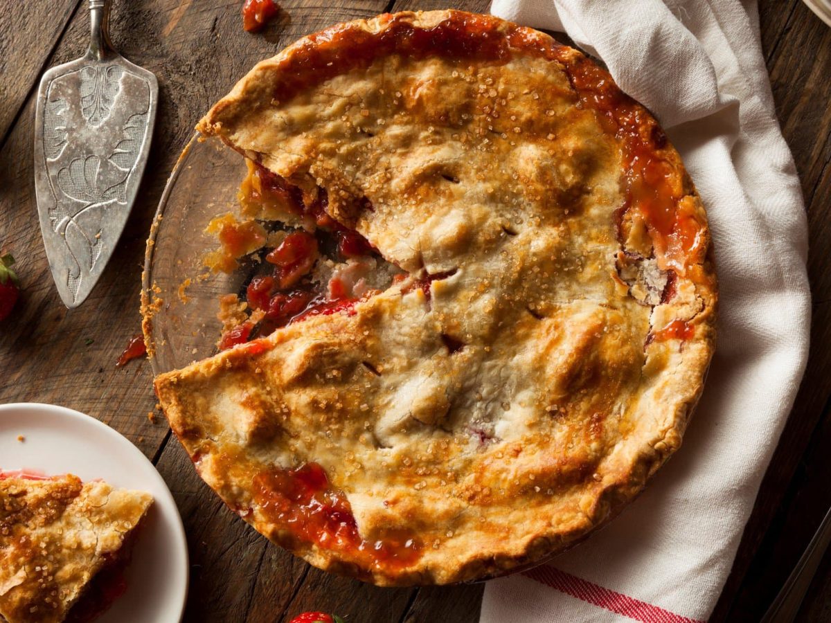 Ein Rhabarber-Pie auf einem Holztisch mit Erdbeeren und einem Tuch. Unten links im Bild noch ein kleiner Teller, mit einem Stück vom Pie.