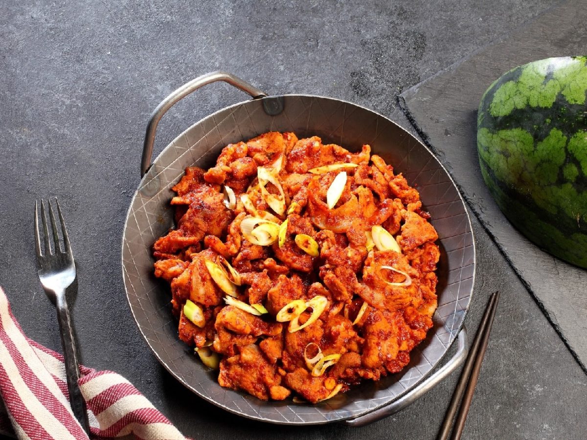 Eine gusseiserne Pfanne mit dem Rindfleisch-Melonen-Ragout auf dunklem Untergrund. Unten links im Bild ein Tuch und oben rechts im Bild eine Wassermelonenspalte.