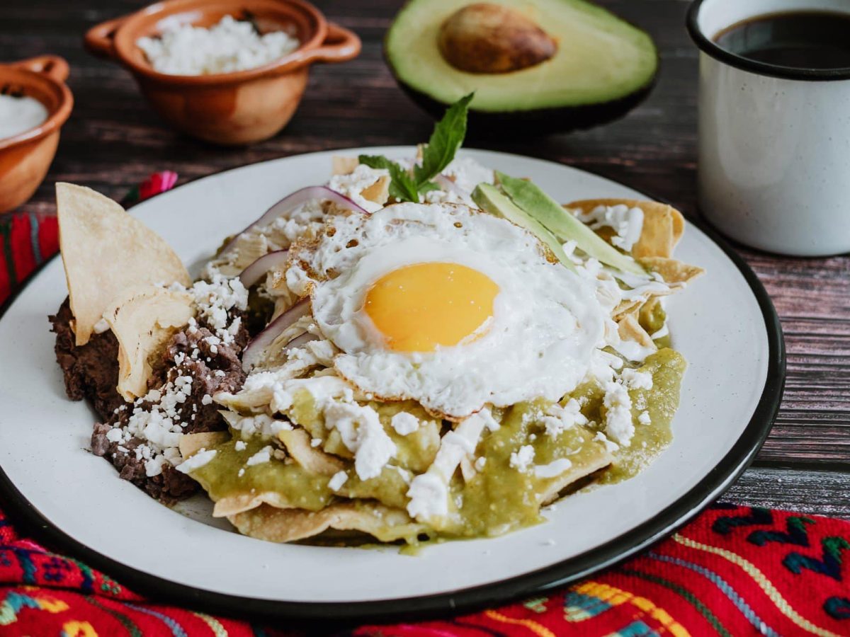 Salsa Verde Baked Eggs auf weißem Teller und grauem Untergrund. Daneben folkloristisches Tuch, Schalen mit geriebenem weißem Käse, eine halbe Avocado und eine Tasse Kaffee.