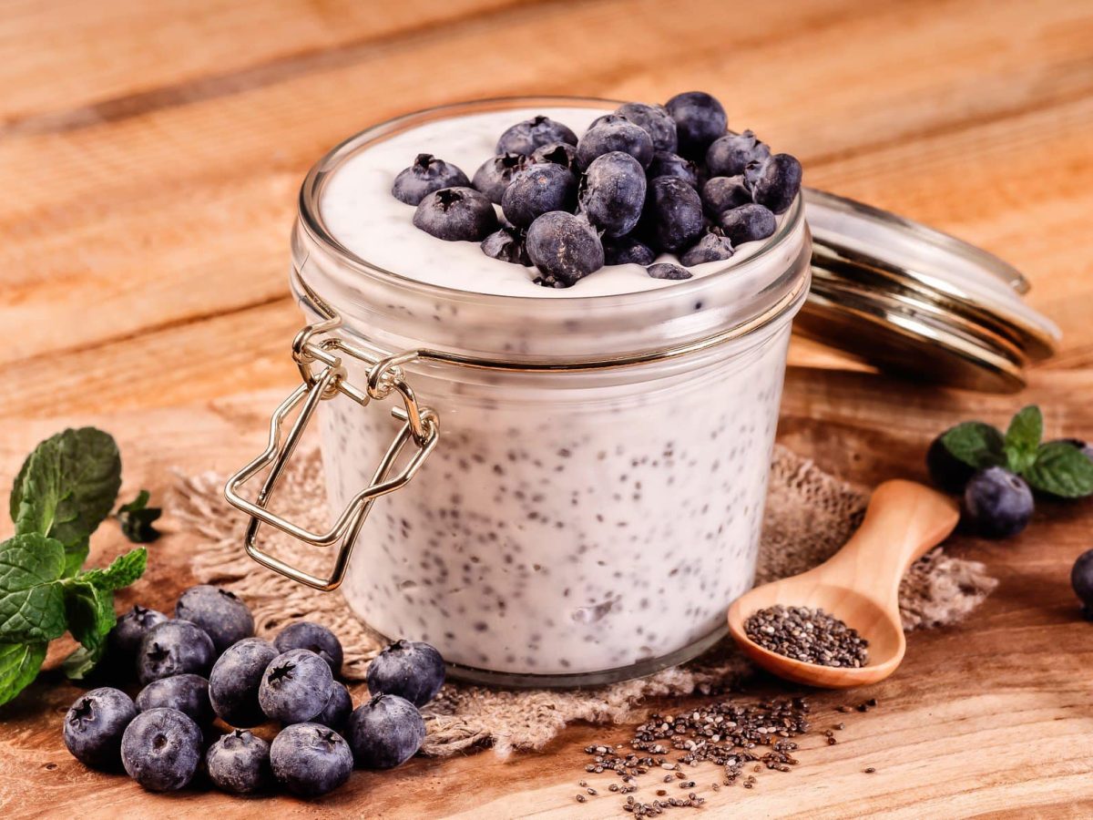 Schnelle Frühstücksbowl mit Blaubeeren und Chiasamen in Einmachglas. Daneben Minze, Blaubeeren und Chiasamen auf Holztisch. Rechts Holzlöffel. Frontalansicht.