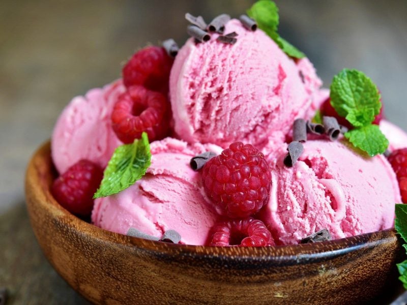 Blitzschnelles Himbeer-Joghurt-Eis mit frischen Himbeeren, Minze und Schokoladenflocken in Tonschale auf hellem Holztisch. Frontalansicht.