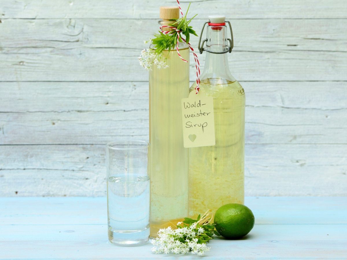 Selbst gemachter Waldmeister-Sirup in zwei Flaschen auf blauem Untergrund vor hellem Holz. An der rechten Schild mit der Aufschrift Waldmeister-Sirup. Frischer Waldmeister als Dekoration an den Flaschen und im Bild. Frontalansicht.