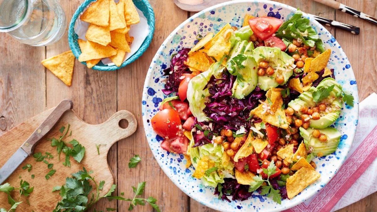 Eine Taco-Bowl in einem hellblau-weißen Teller auf einem Holztisch neben einem Holzschneidebrett und frischer Petersilie