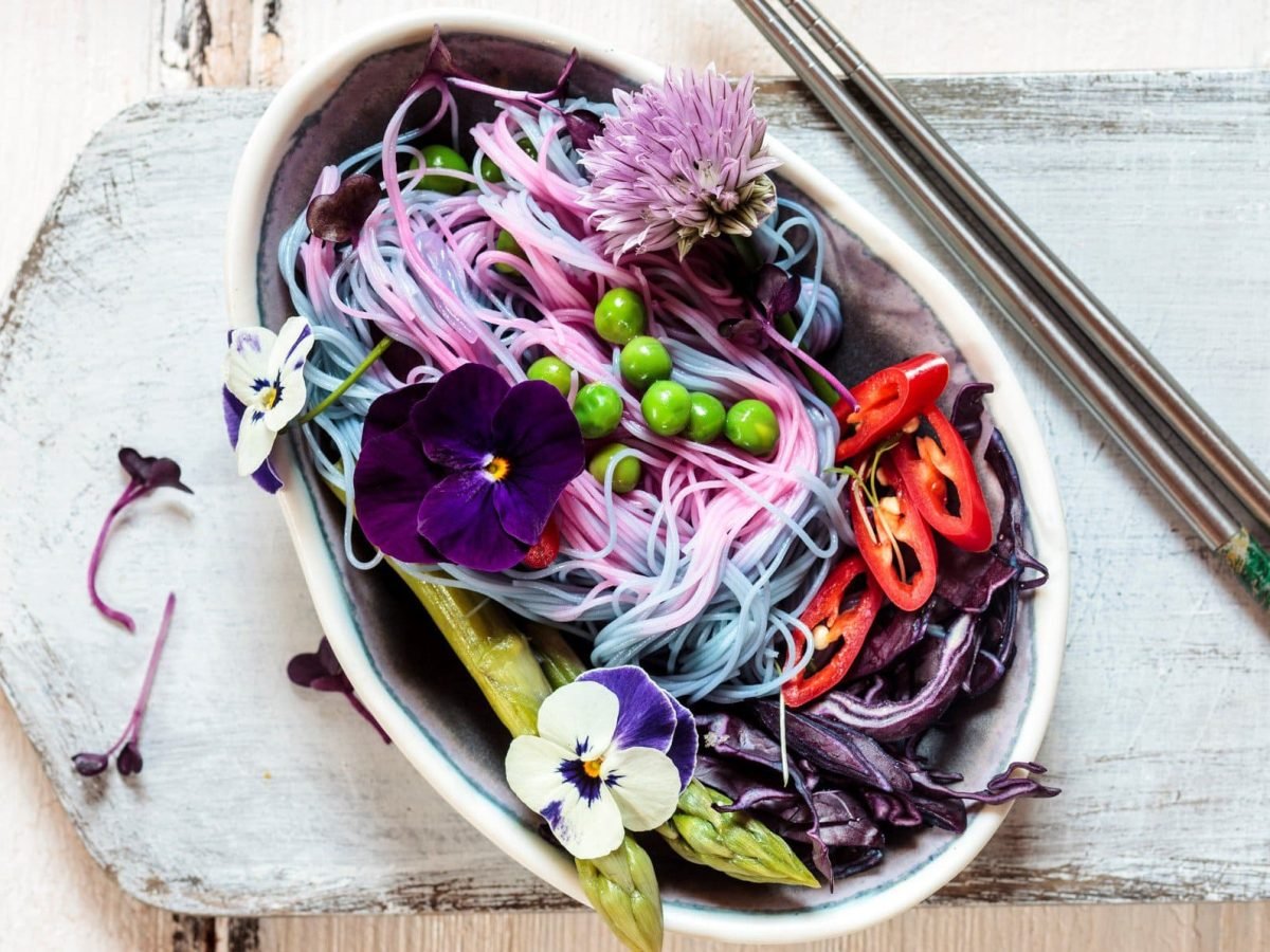 Eine ovale Schale mit den Unicorn Noodles und verschiedenem Gemüse wie Spargel, Rotkohl, Erbsen und dekorativen Blüten.