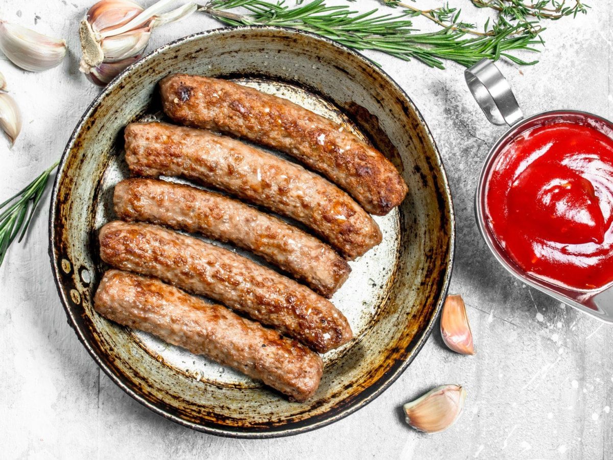 Vegane Bratwürste auf einem Teller, daneben Knoblauch und Kräuter sowie ein Kännchen rote Grillsauce, in der Draufsicht.