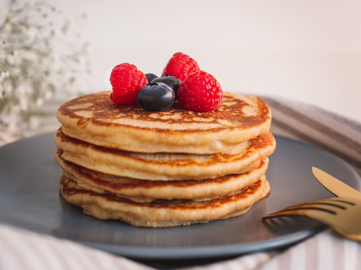 Ein Stapel vegane Pancakes mit frischen Beeren auf einem dunkelgrauen Teller, daneben goldenes Besteck.