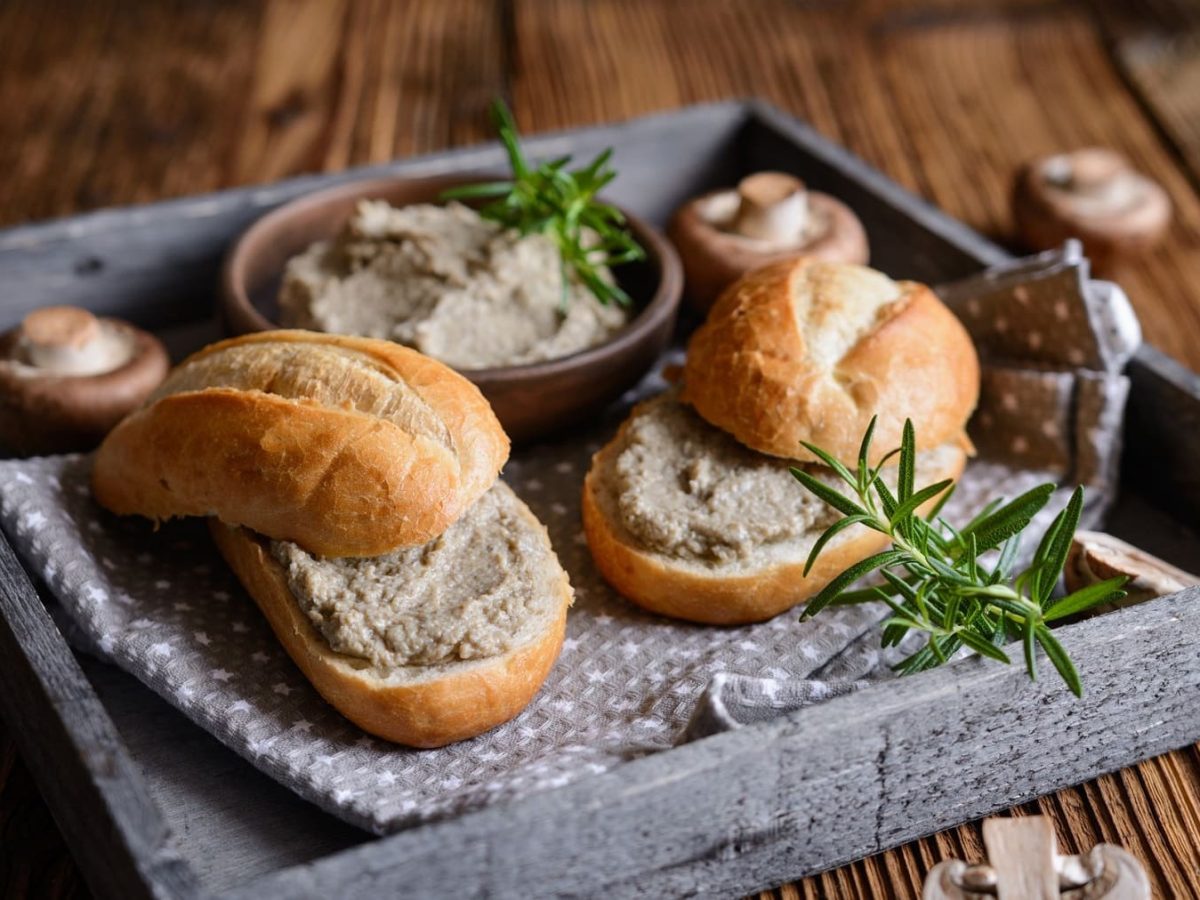 Zwei Brötchen mit veganem Brotaufstrich mit Chamoignons auf einem Tablett, außerdem eine Schale mit dem Brotaufstrich und frische Pilze.