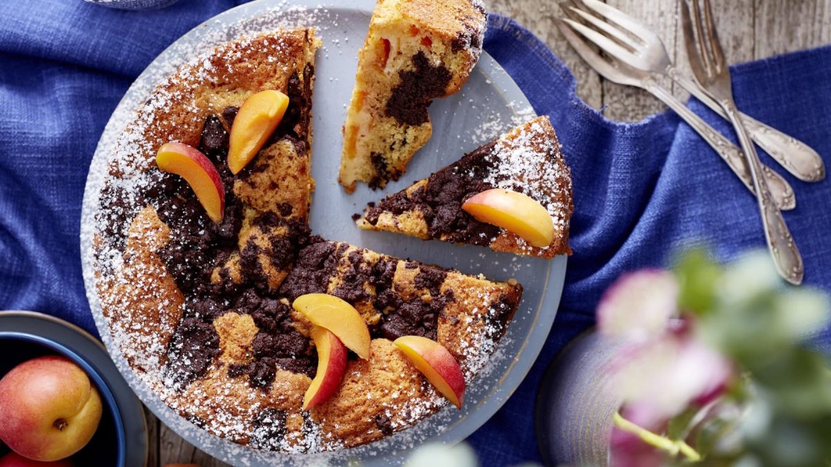 Wolkenkuchen mit Schokostreuseln auf einem Teller. Daneben ein Strauß Blumen, Besteck und eine Aprikose.