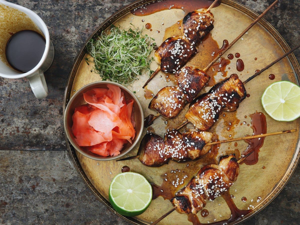 Gegrillte Yakitori japanische Hähnchenspieße mit Sesam, Limette, Kresse und einer Schale eingelegtem Ingwer auf beigem Tonteller. Daneben weiße Kanne mit Ponzu-Sauce auf dunklem Holz. Aufnahme aus der Vogelperspektive.