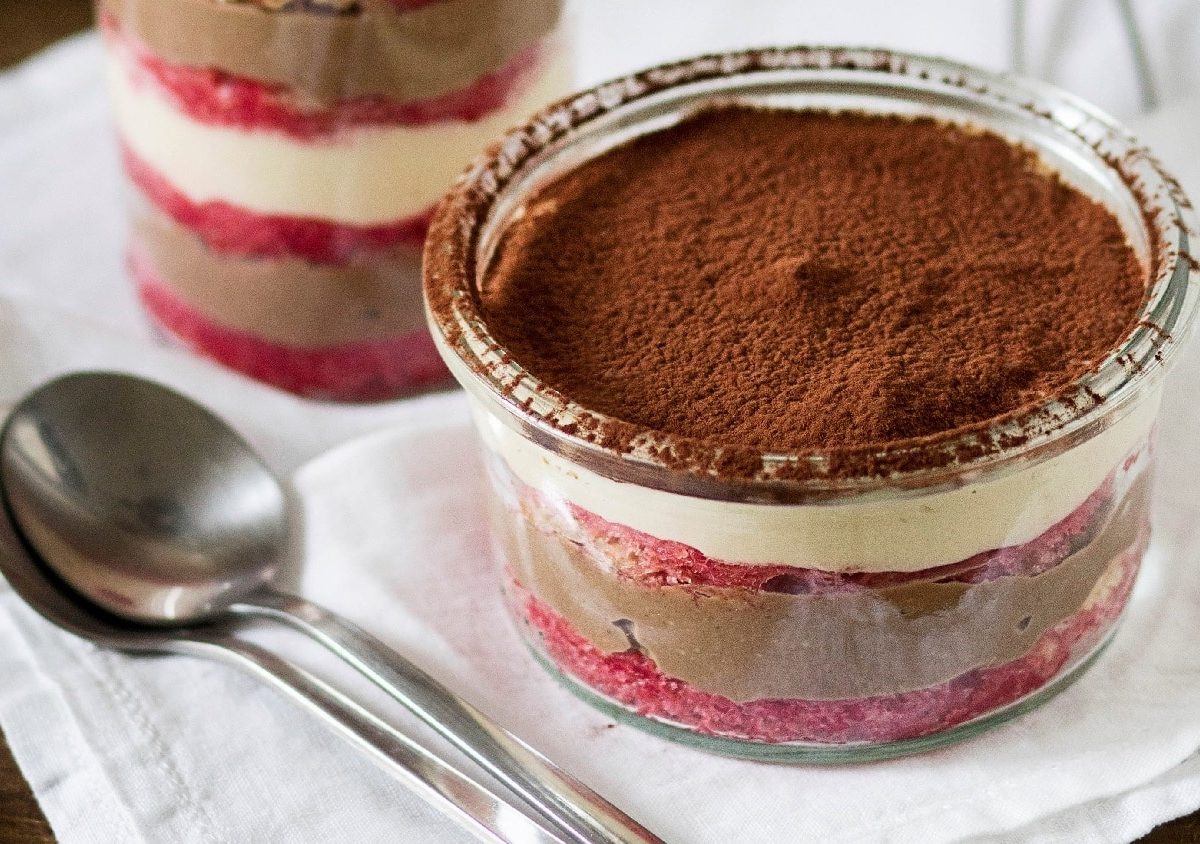 Zwei Gläser des Schichtdesserts auf einer weißen Tischdecke mit zwei silbernen Löffeln im Vordergrund.