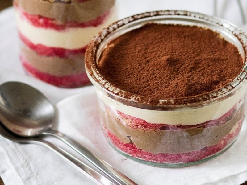 Zwei Gläser des Schichtdesserts auf einer weißen Tischdecke mit zwei silbernen Löffeln im Vordergrund.