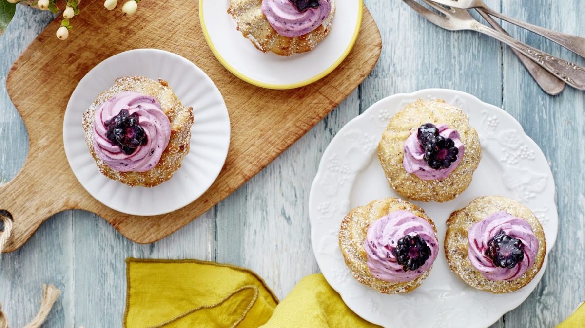 Auf einem Teller stehen 3 Apfelmus-Brombeer-Gugelhupf. Sie sind mit einer lila Creme und je einer Brombeere verziert.